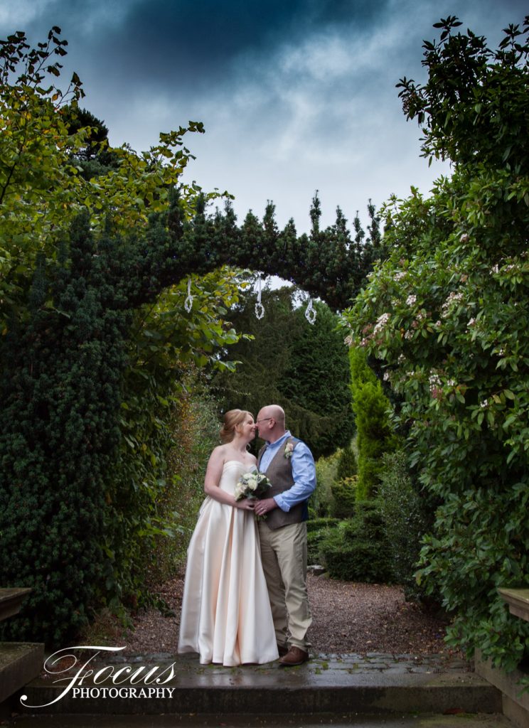 A bride and groom stand under a leafy archway in a garden, kissing and holding a bunch of flowers, with cloudy skies overhead. Focus Photography by Tim Teasdale