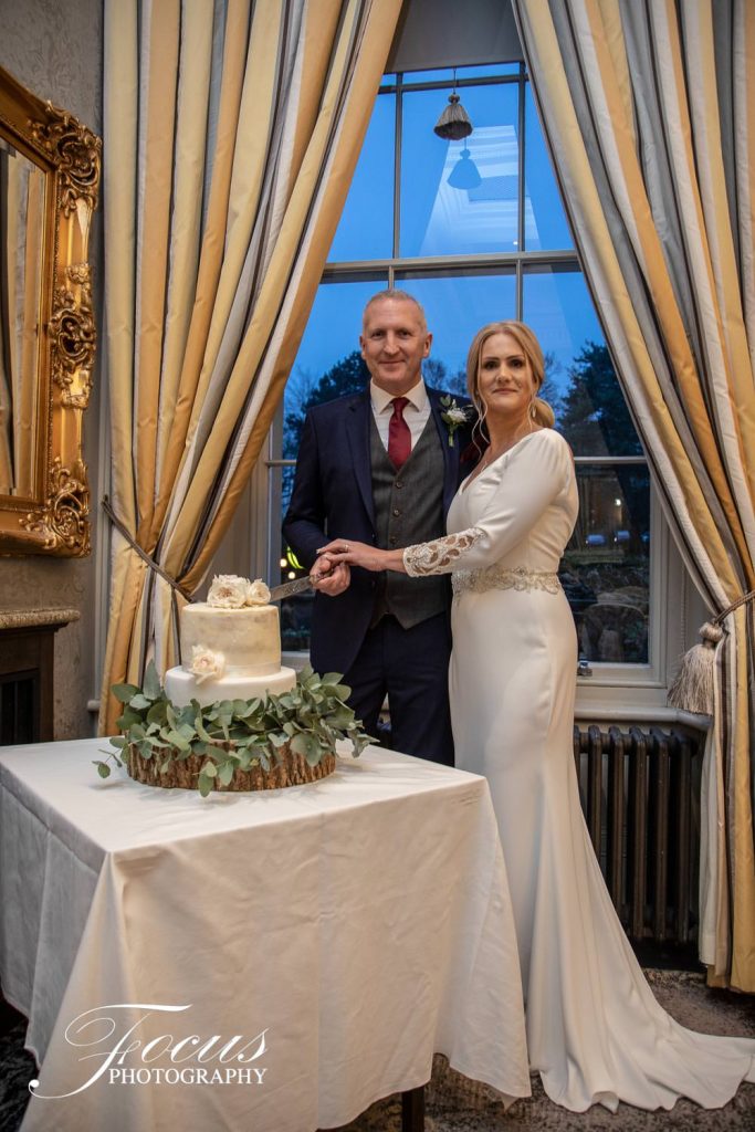 A bride and groom stand together indoors beside a decorated wedding cake, posing for a photo in front of tall windows with curtains. Focus Photography by Tim Teasdale