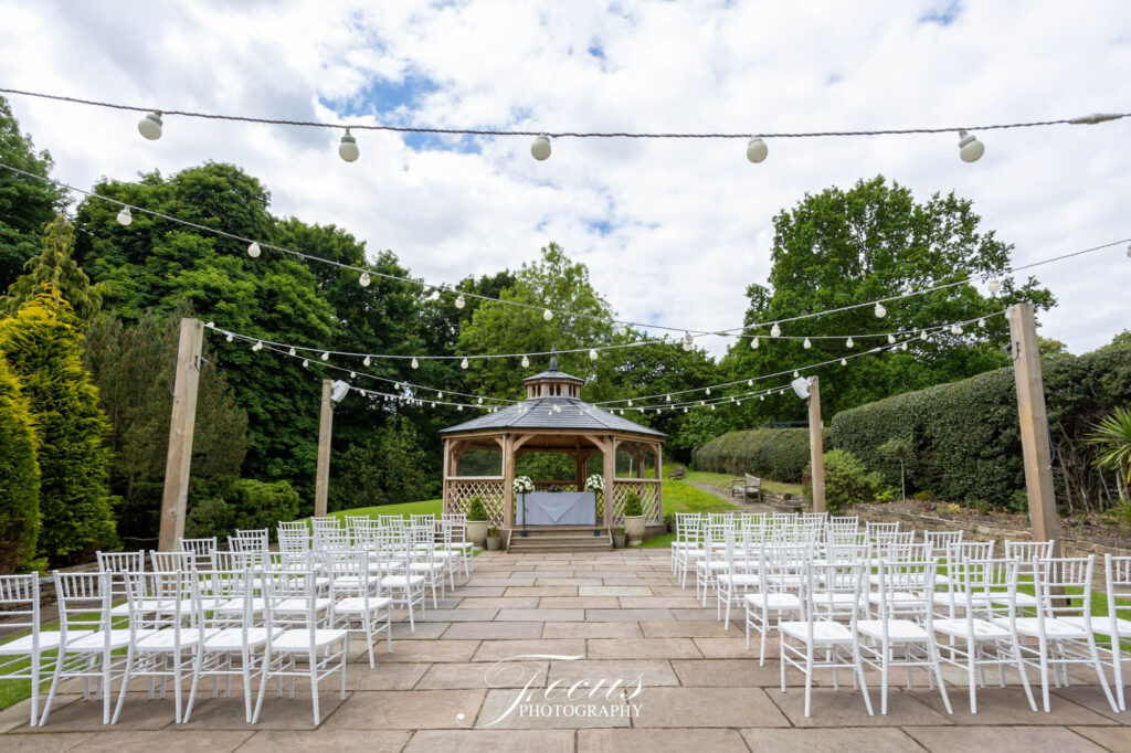 Outdoor wedding ceremony setup with rows of white chairs facing a wooden gazebo, fairy lights overhead, and greenery in the background