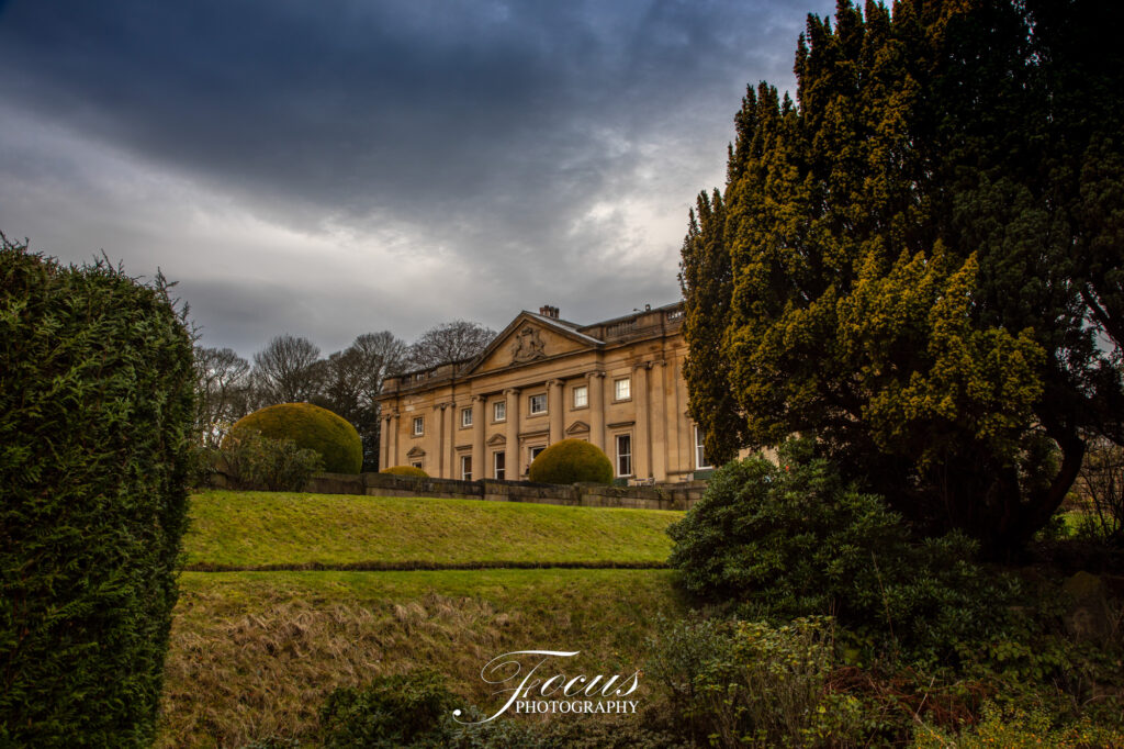 A large, historic stone mansion stands on a grassy hill, surrounded by trimmed bushes and tall trees under a cloudy sky