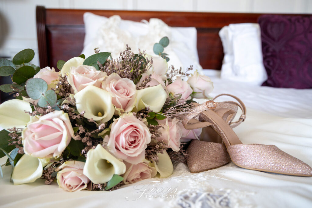 A bouquet of pale pink roses and white calla lilies rests next to a pair of sparkly, blush-coloured shoes on a neatly made bed