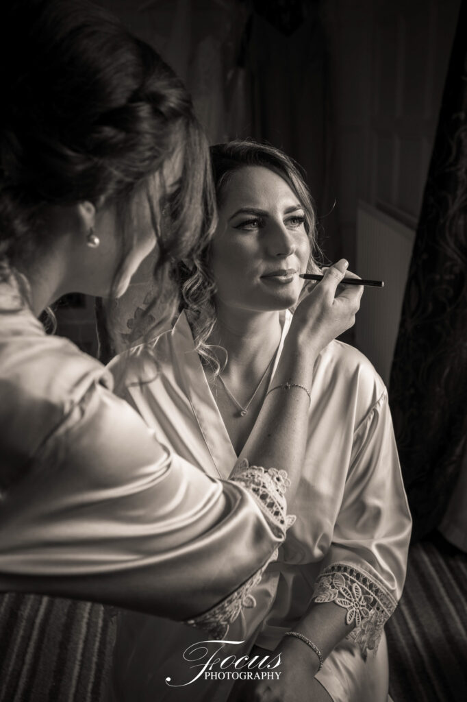 A woman applies make-up to another woman in a satin dressing gown, who is seated and facing forwards, in a softly lit room