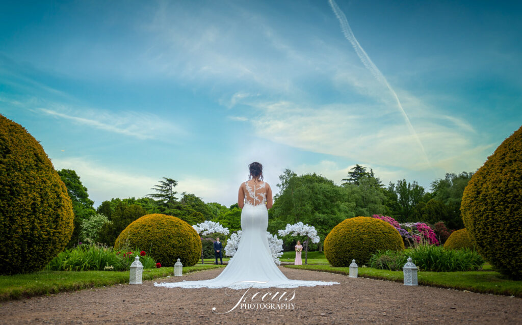 A bride in a white wedding dress stands on a garden path, facing away, surrounded by neatly trimmed bushes and lanterns, with guests visible in the distance