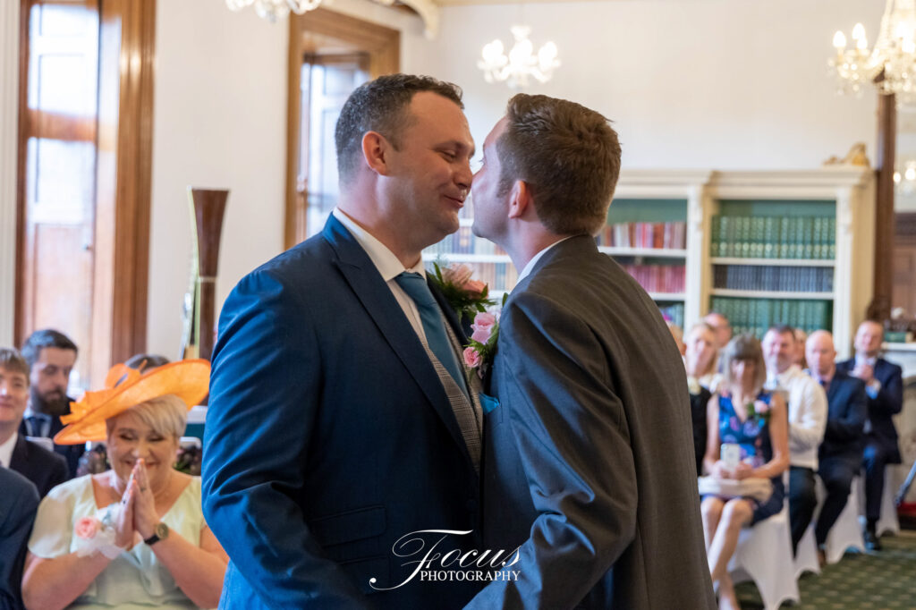 Two men in suits share a moment at the front of a room during a wedding ceremony, with seated guests watching and applauding in the background