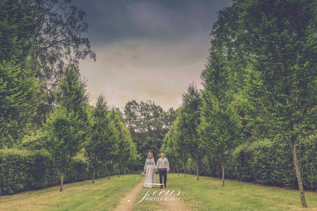 A couple walks hand in hand down a tree-lined path in a park under an overcast sky, with green foliage surrounding them