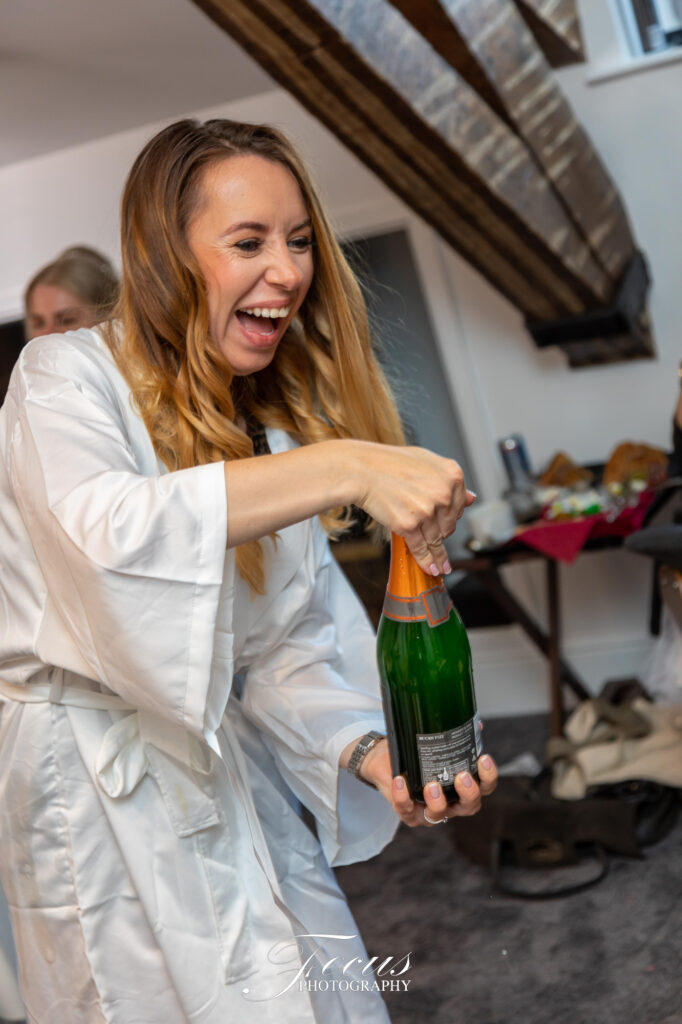 A woman in a white dressing gown laughs whilst opening a bottle of champagne in a room with exposed wooden beams and a table in the background