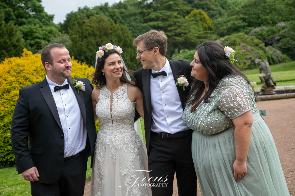 Four adults dressed formally, including a bride and groom, stand together outdoors, smiling and looking at each other on a path with greenery and a statue in the background