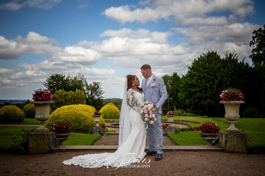 A bride and groom stand together in a garden, facing each other and smiling, surrounded by greenery and flower arrangements under a partly cloudy sky