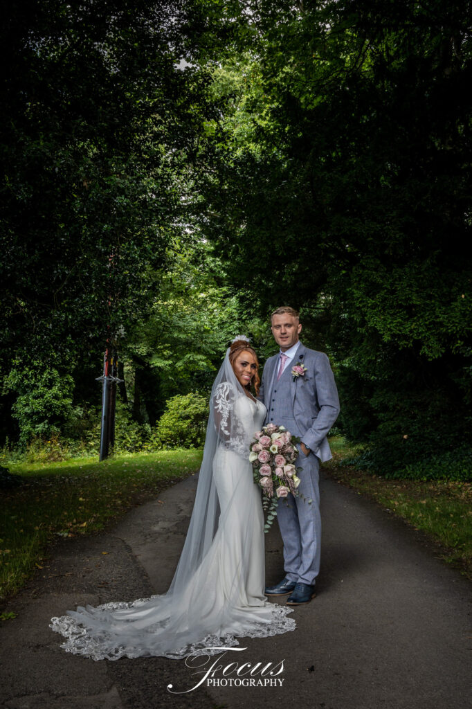 A bride and groom stand together on a paved path surrounded by trees; the bride holds a bouquet of pink and white flowers
