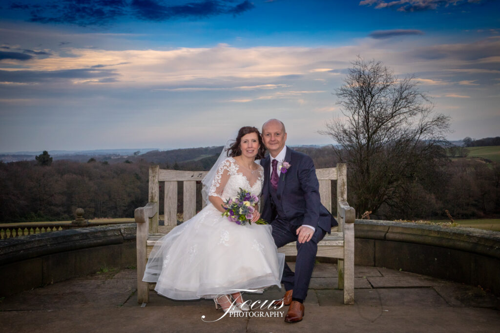 A bride and groom in formal attire sit together on a wooden bench outdoors with a scenic landscape and cloudy sky in the background
