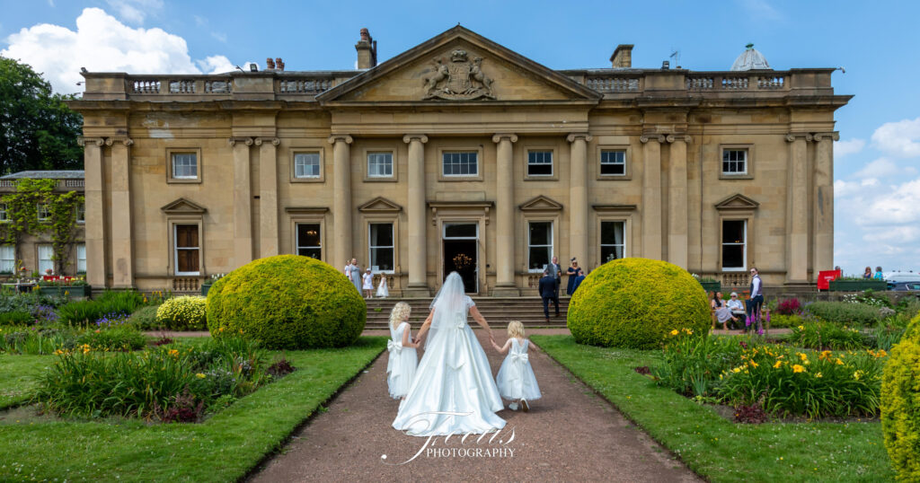 A bride and two young girls in white dresses walk towards a grand stone manor with manicured gardens and guests gathered outside. Focus Photography by Tim Teasdale