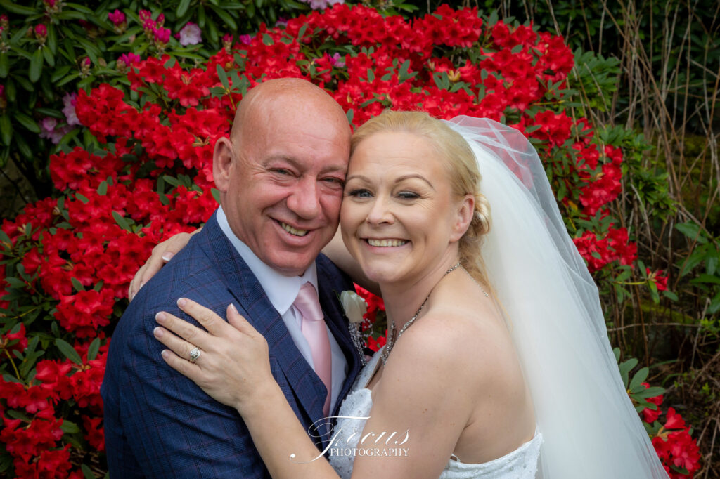 A bride and groom smile and embrace in front of bright red flowers, both wearing wedding attire