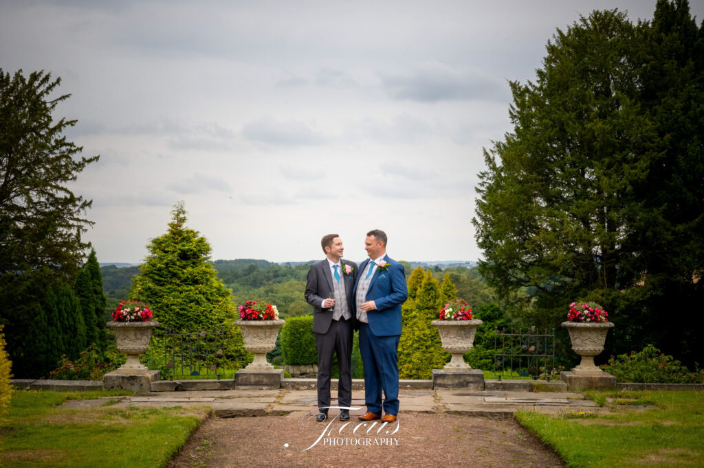Two men in suits stand facing each other, holding hands, on a stone terrace with flower pots and trees in the background