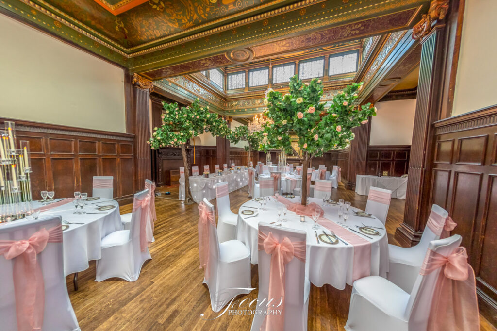 Elegant banqueting hall with decorated round tables, white tablecloths, pink chair sashes, floral centrepieces, and ornate wood panelling and ceiling