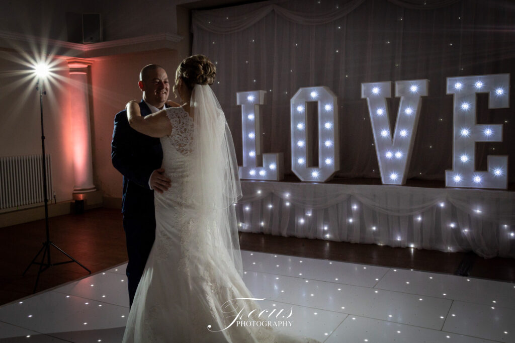 A bride and groom share a first dance in a dimly lit room with large illuminated letters spelling "LOVE" in the background