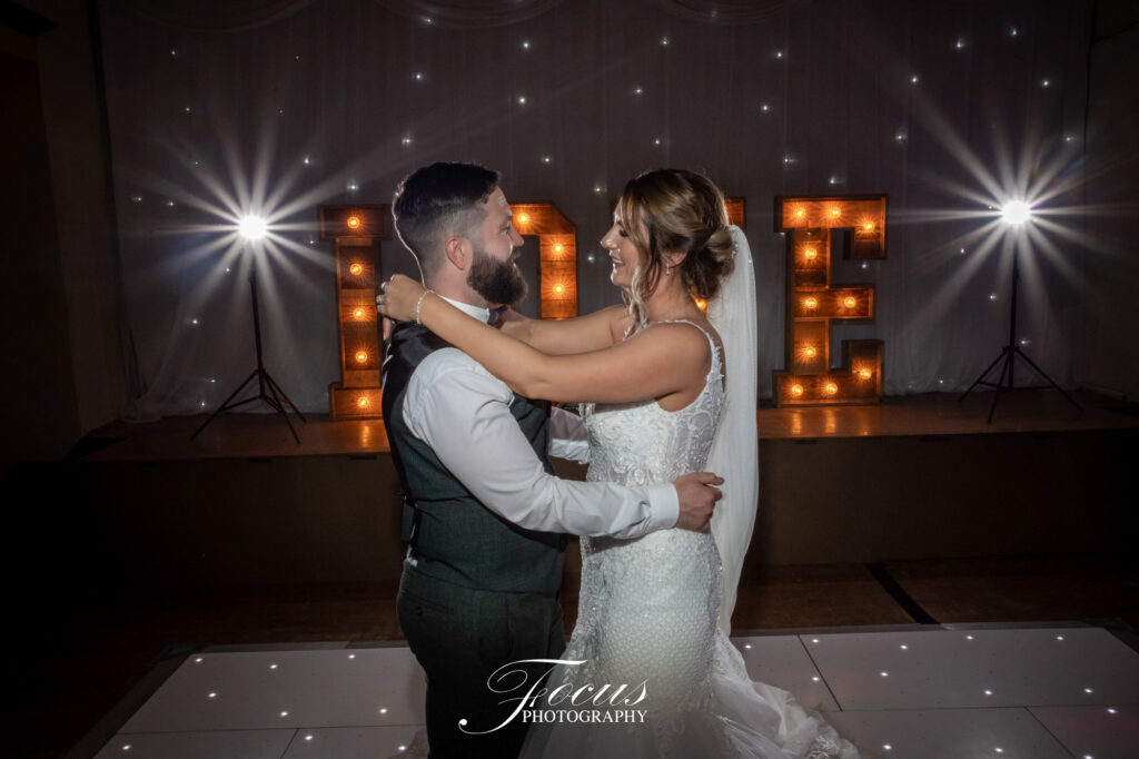 A bride and groom dance together on a white-lit floor with large illuminated "LOVE" letters and spotlights in the background