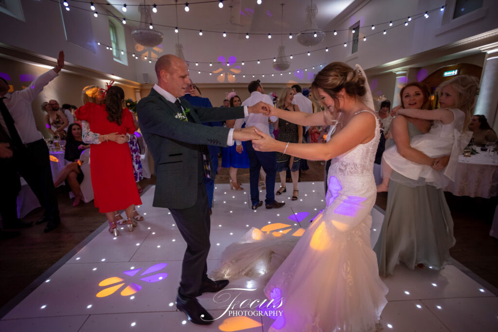 A bride and groom dance together on a lit, decorated floor at their wedding reception, surrounded by guests in a decorated hall