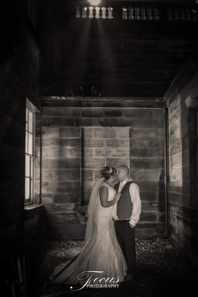 A bride and groom share a kiss in a dimly lit stone corridor, with a beam of light shining down from above
