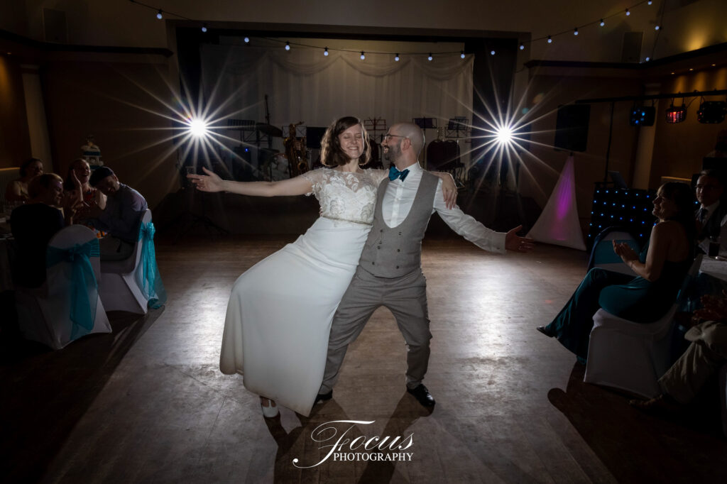 A bride and groom pose playfully on the dance floor at their wedding reception, smiling, with guests seated around the room and bright lights in the background