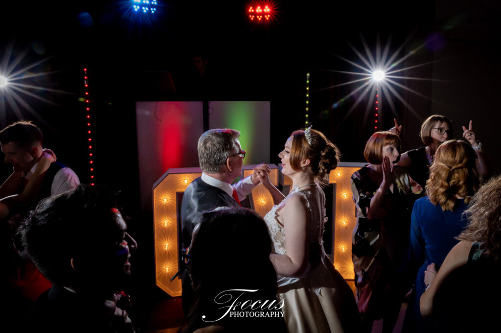 A bride and groom dance at their wedding reception, surrounded by guests, colourful lights, and large illuminated letters, with a DJ booth in the background