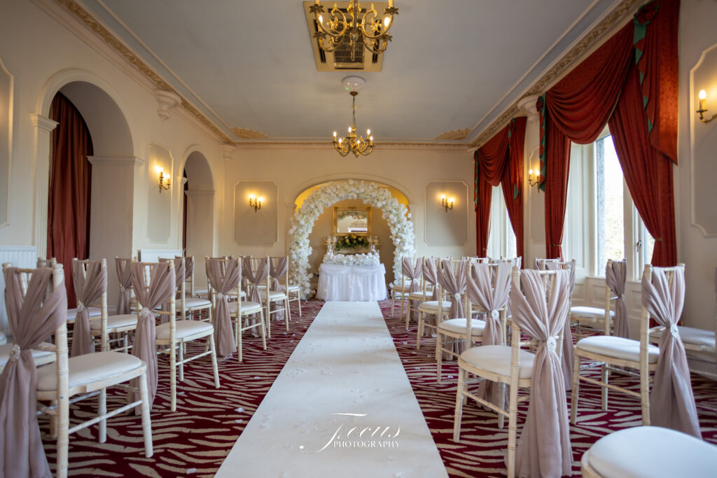 A wedding ceremony room with rows of white chairs, taupe chair covers, a white aisle runner, floral arch, and red curtains. Focus Photography by Tim Teasdale