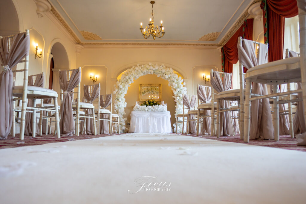 Rows of chairs with beige sashes face a white floral arch and table in an elegantly decorated indoor wedding ceremony venue. Focus Photography by Tim Teasdale