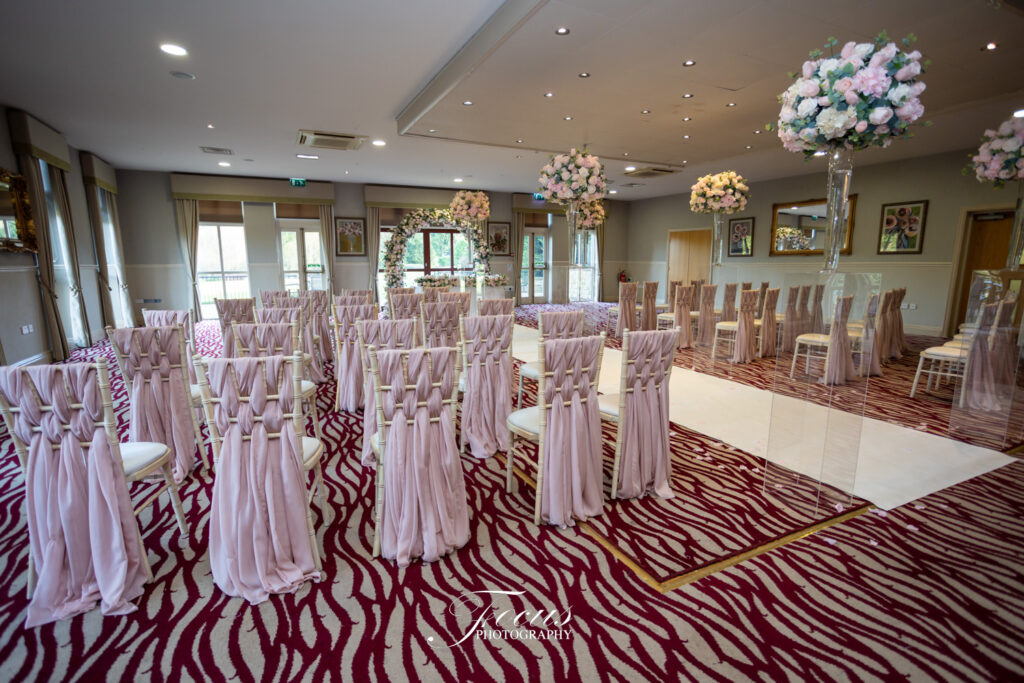 A wedding ceremony room with rows of chairs draped in light pink fabric, floral arrangements on tall stands, and a white carpet aisle. Focus Photography by Tim Teasdale
