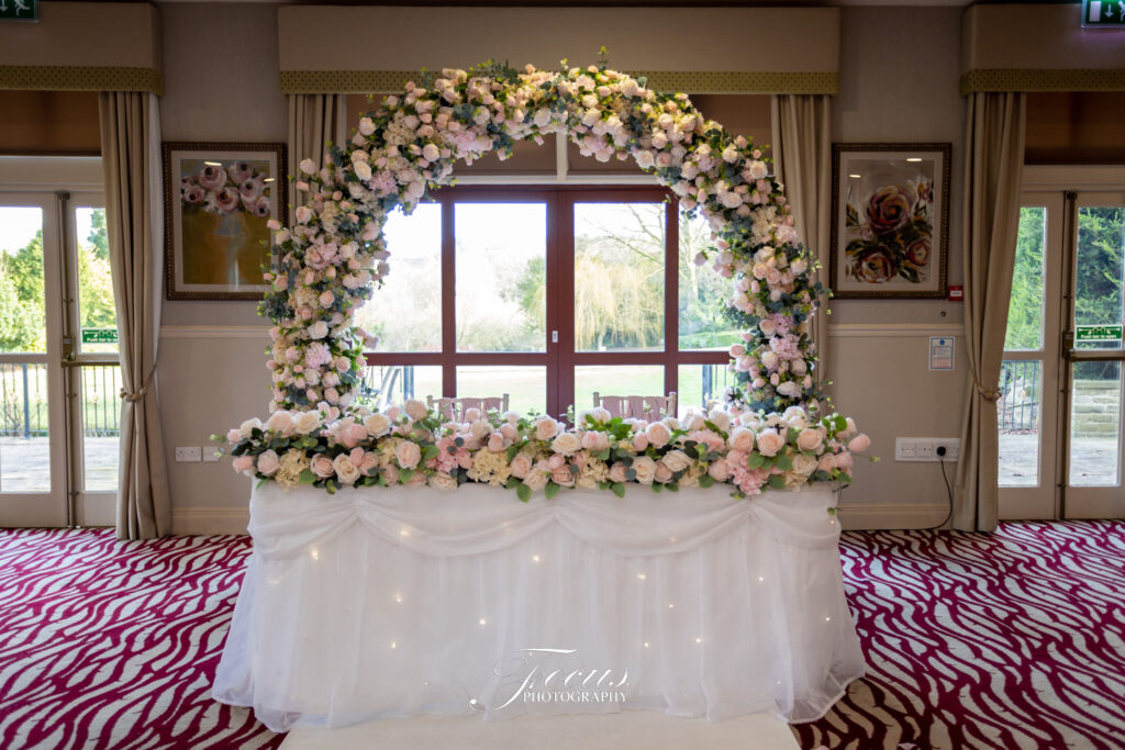 A decorated wedding table with a white tablecloth and an arch of pink and white flowers, set in front of large windows in an indoor venue. Focus Photography by Tim Teasdale