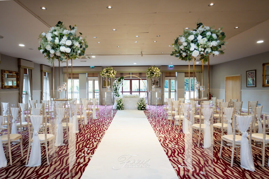 A decorated indoor wedding ceremony set-up with rows of chairs, tall floral arrangements, and a white aisle runner leading to an altar. Focus Photography by Tim Teasdale