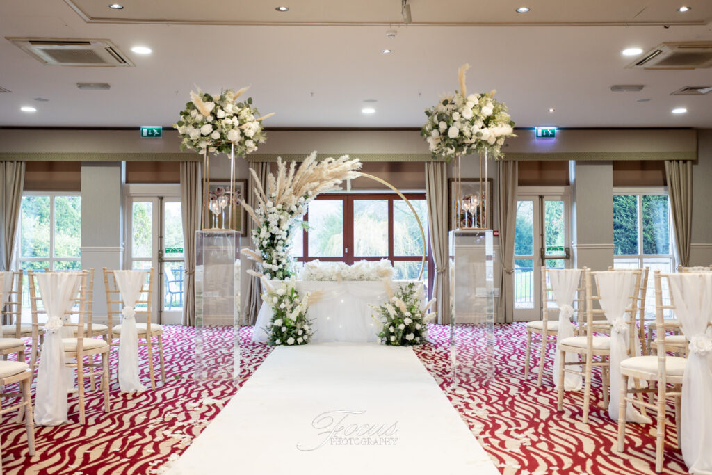 Indoor wedding ceremony set-up with white chairs, floral arrangements, and a circular arch at the front, arranged on a white aisle runner over a patterned red carpet. Focus Photography by Tim Teasdale