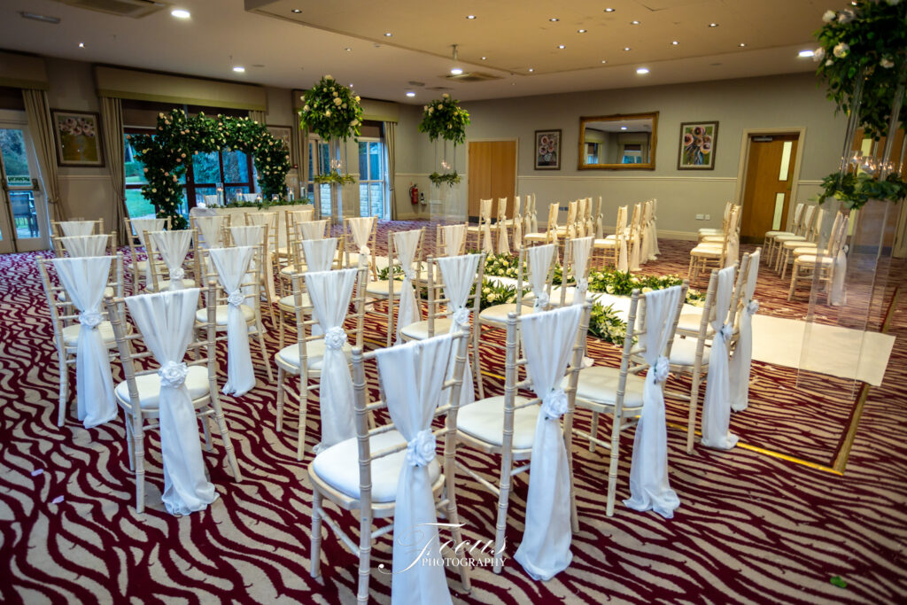 Rows of white chairs with white fabric decorations are arranged indoors for a wedding ceremony, with floral arrangements and a patterned carpet visible. Focus Photography by Tim Teasdale