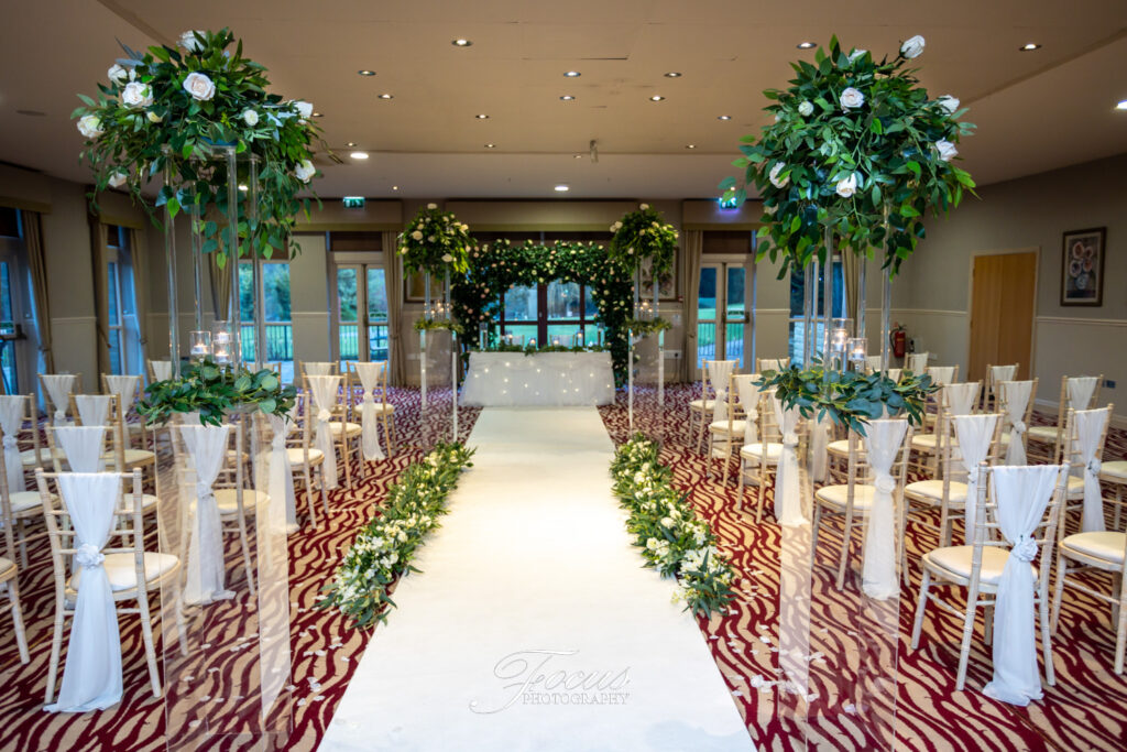 A decorated indoor wedding aisle with white chairs, greenery, floral arrangements, and an altar at the front under a canopy of lights. Focus Photography by Tim Teasdale