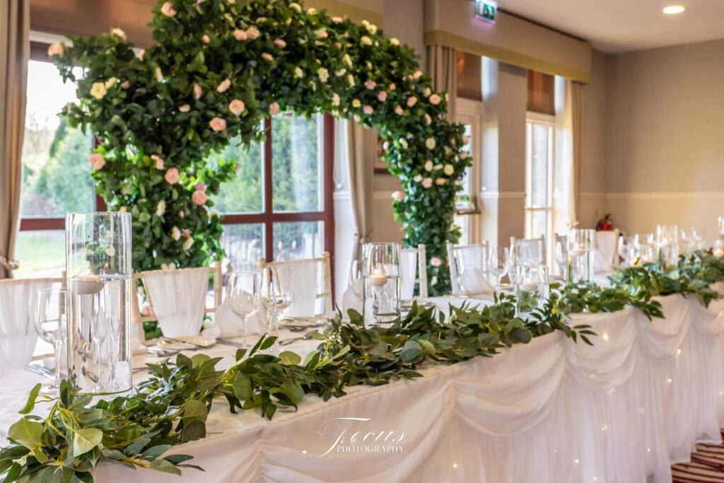 A long banquet table decorated with white linen, greenery, candles, and glassware, set in front of a floral arch in a bright, elegant room. Focus Photography by Tim Teasdale
