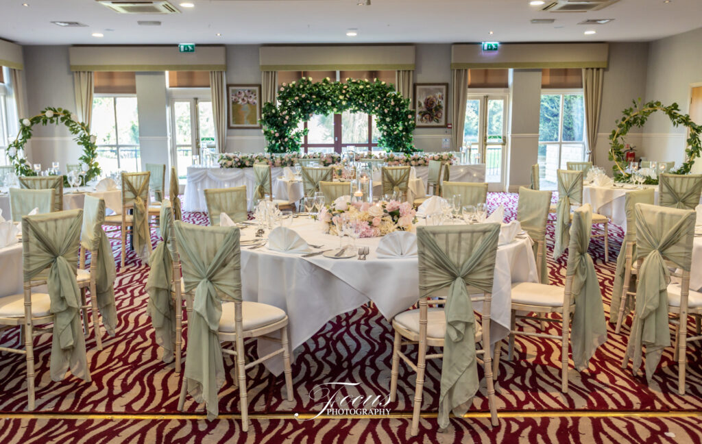 A wedding reception venue with round tables, white tablecloths, green chair sashes, floral centrepieces, and a decorated greenery arch at the front. Focus Photography by Tim Teasdale