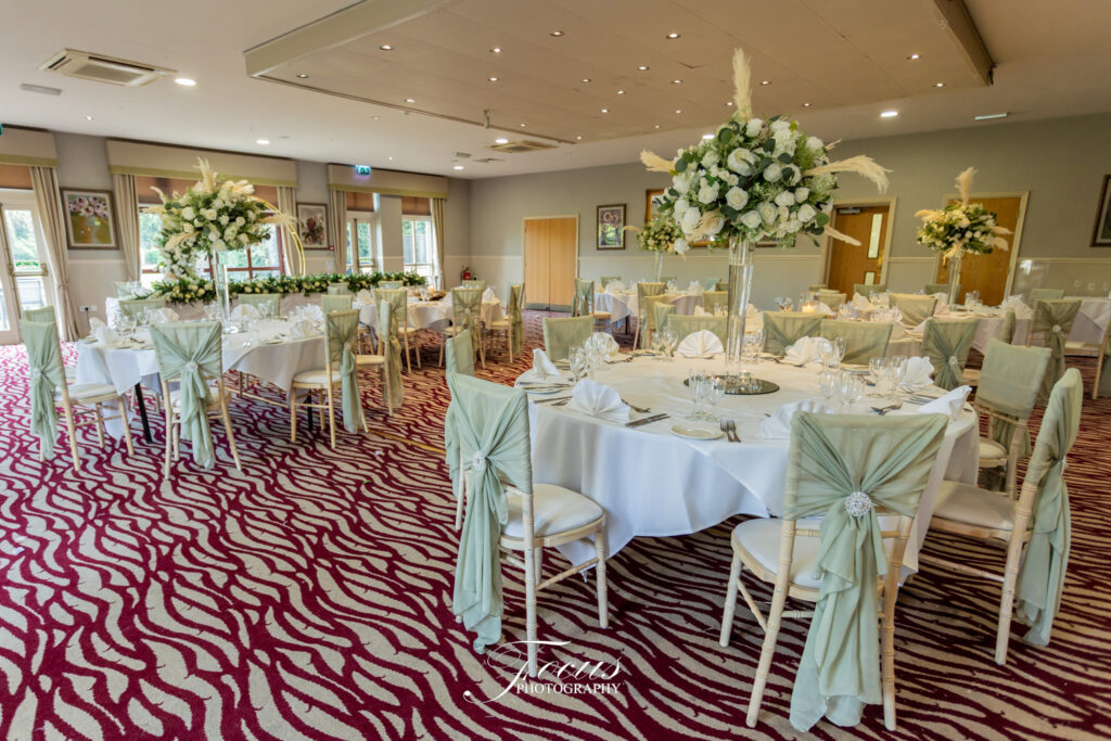 A banqueting hall with round tables set for an event, featuring white tablecloths, floral centrepieces, and chairs decorated with sage green bows. Focus Photography by Tim Teasdale