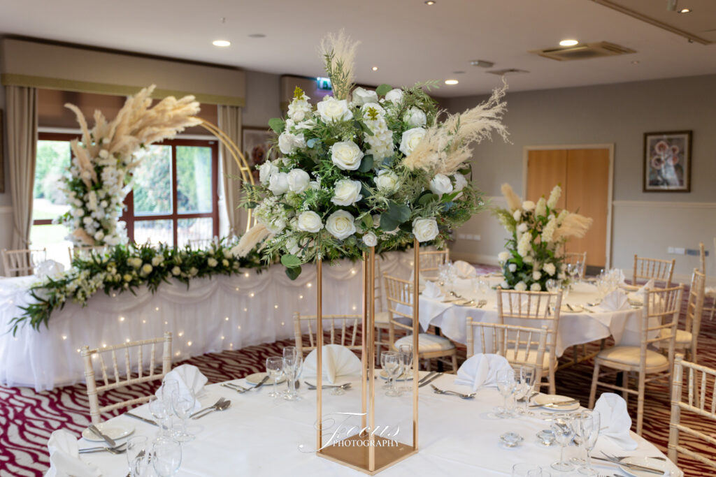 Round table set for an event with a tall centrepiece of white roses and greenery; decorated room with white flowers and chairs arranged neatly. Focus Photography by Tim Teasdale