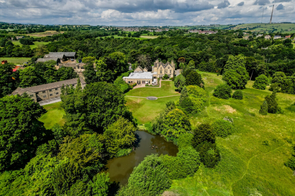 Aerial view of a large country house surrounded by trees, gardens, and a pond, with fields and distant buildings in the background under a partly cloudy sky. Focus Photography by Tim Teasdale