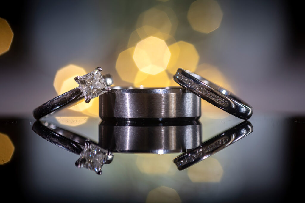 Three silver rings, including one with a square-cut diamond, are displayed on a reflective surface with blurred yellow lights in the background. Focus Photography by Tim Teasdale