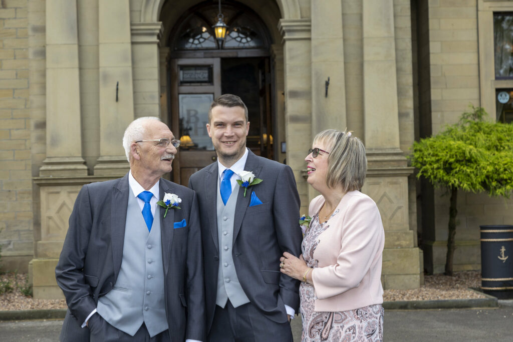 Three people dressed formally stand smiling in front of a stone building entrance, with two men in suits and a woman in a light pink outfit. Focus Photography by Tim Teasdale