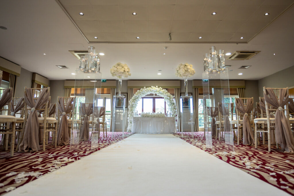 Indoor wedding ceremony setup with a white aisle runner, flower arrangements, and chairs draped in taupe fabric, facing a floral arch and table at the front of the room. Focus Photography by Tim Teasdale