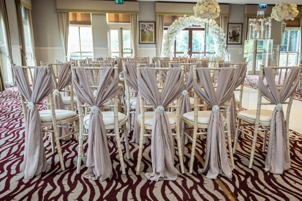 Rows of white chairs with pale fabric sashes are arranged for a wedding ceremony in a decorated indoor venue with an arch and floral displays. Focus Photography by Tim Teasdale