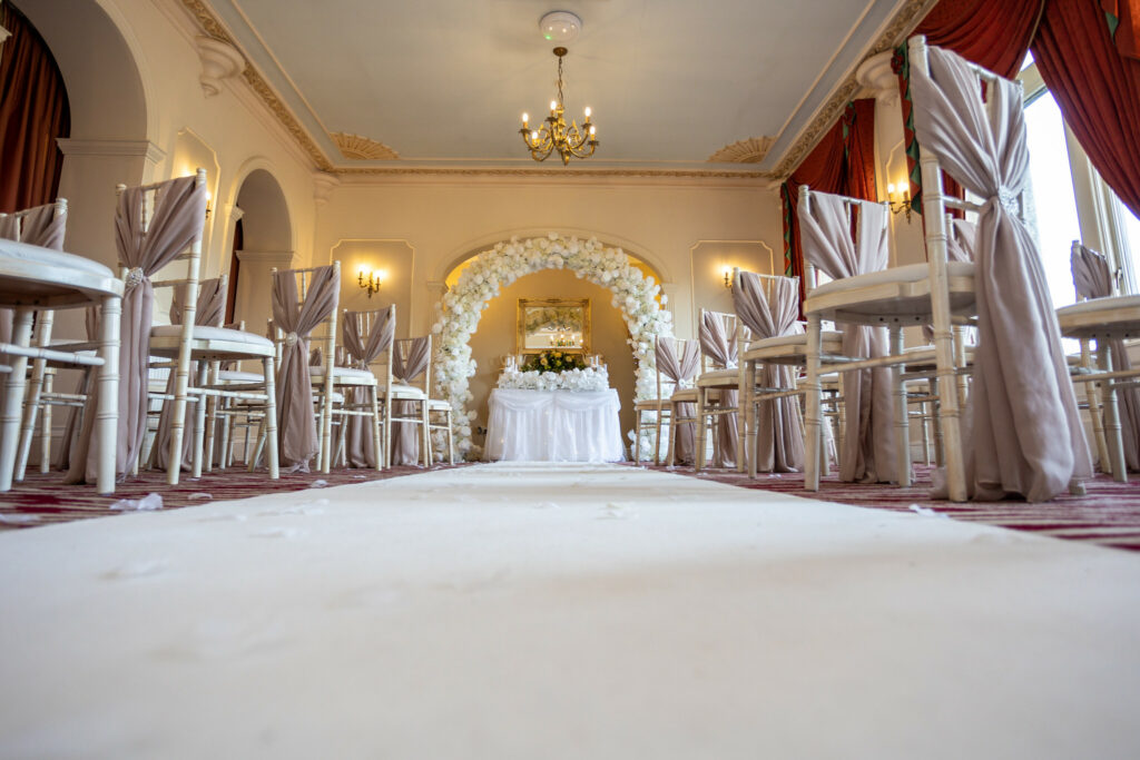 A wedding ceremony set-up with decorated chairs, a white aisle runner, and a floral arch in an elegant room with chandeliers and red curtains. Focus Photography by Tim Teasdale
