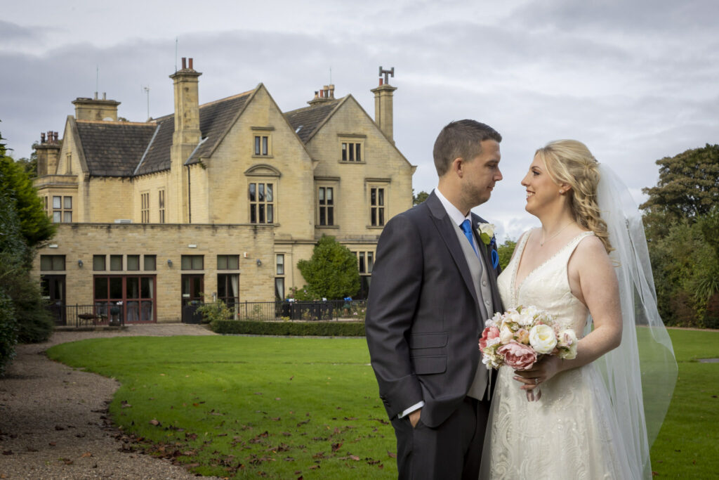 A bride and groom stand together outside a large stone building, facing each other and holding a bunch of flowers. Focus Photography by Tim Teasdale