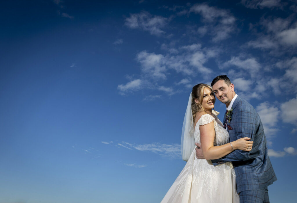 Bride and groom in wedding attire embrace and smile at the camera against a bright blue sky with scattered clouds. Focus Photography by Tim Teasdale
