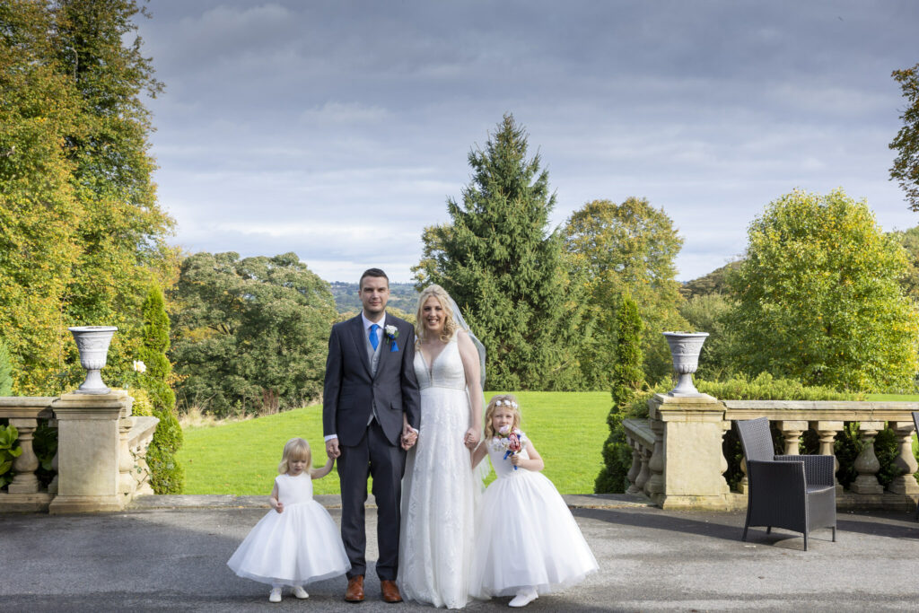 A bride and groom stand outdoors on a stone terrace with two young flower girls in white dresses, surrounded by greenery and trees. Focus Photography by Tim Teasdale