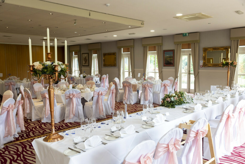 A banqueting hall set for an event with round and rectangular tables, white chairs with pink bows, crockery, floral centrepieces, and candles. Large windows provide natural light. Focus Photography by Tim Teasdale