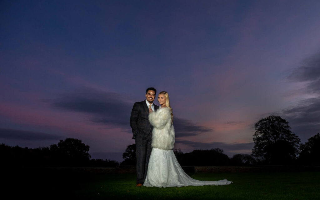 A couple dressed in wedding attire stands on grass at dusk, with a dark sky and silhouettes of trees in the background. Focus Photography by Tim Teasdale