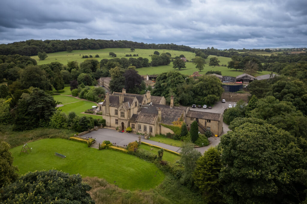 A large stone country house surrounded by trees and fields, with a cloudy sky overhead and a drive leading to the main entrance. Focus Photography by Tim Teasdale