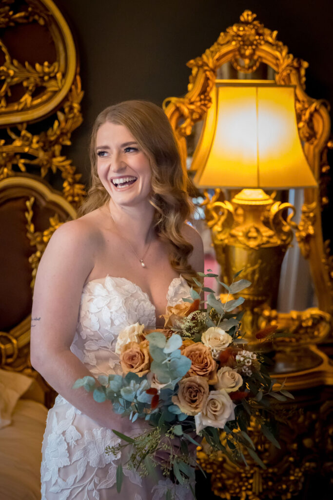 A bride in a strapless white dress holding a bouquet of flowers stands smiling beside an ornate gold mirror and lamp. Focus Photography by Tim Teasdale