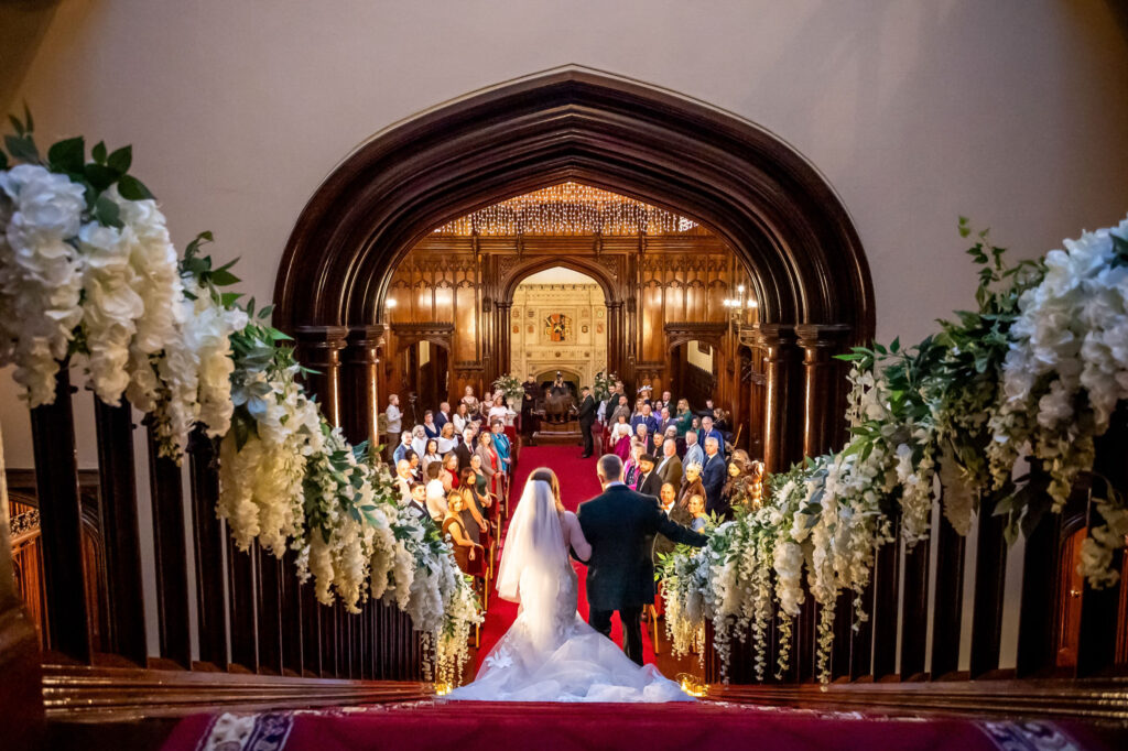 A bride and groom stand at the top of a flower-decorated staircase, facing guests seated in a church or grand hall for their wedding ceremony. Focus Photography by Tim Teasdale